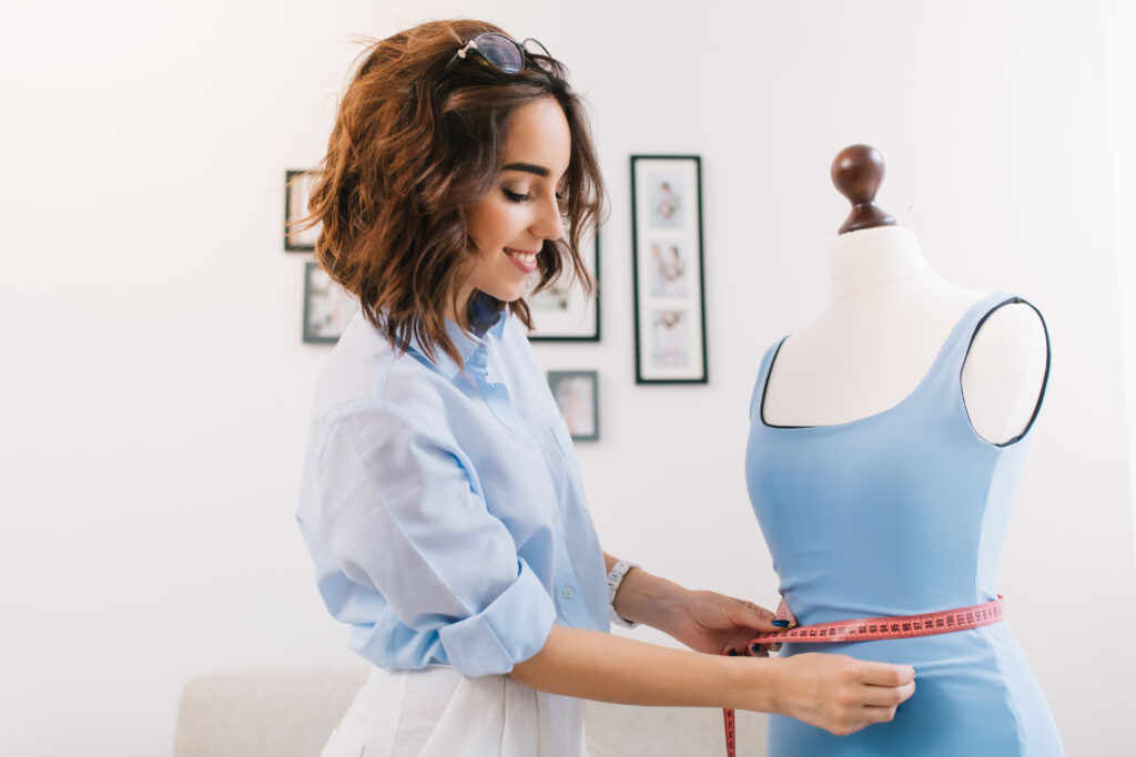 A brunette girl in a blue shirt is working in the workshop studio of making wholesale bodysuits. She makes fitting on the blue dress on the  mannequin.